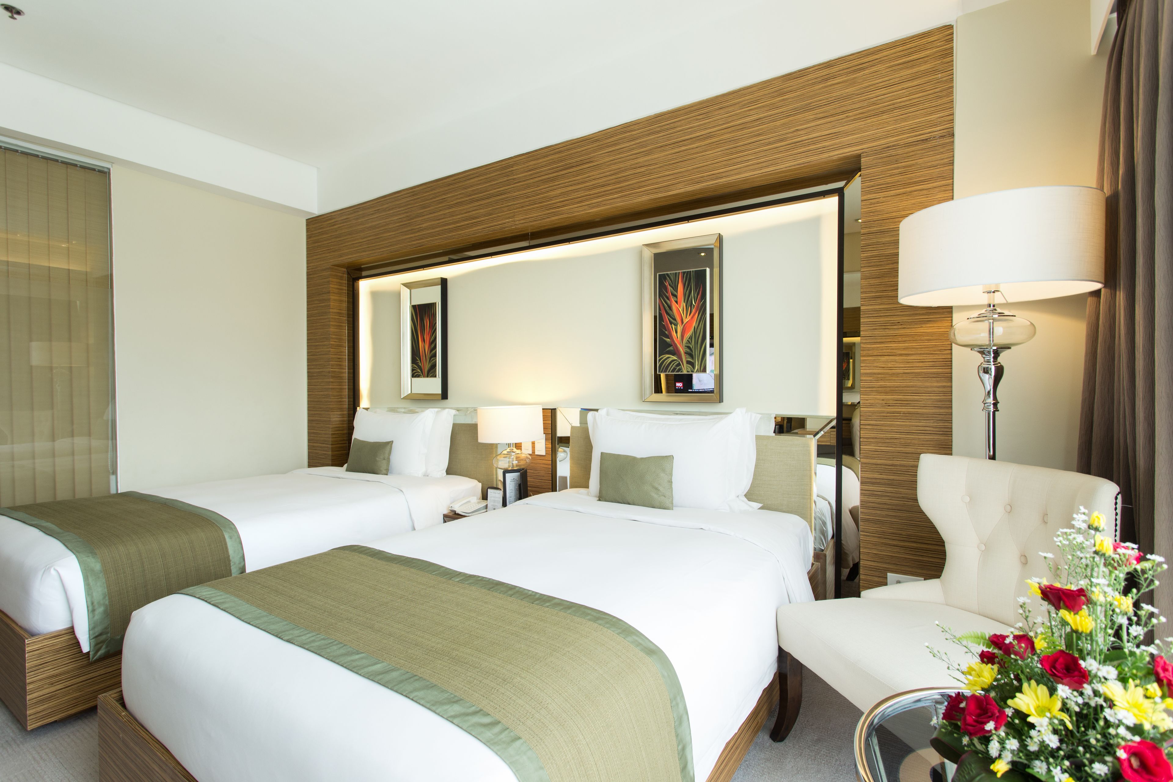 Habitación de hotel con dos camas, una zona de estar con una silla, una mesa con un jarrón de flores y un espejo grande sobre las camas.