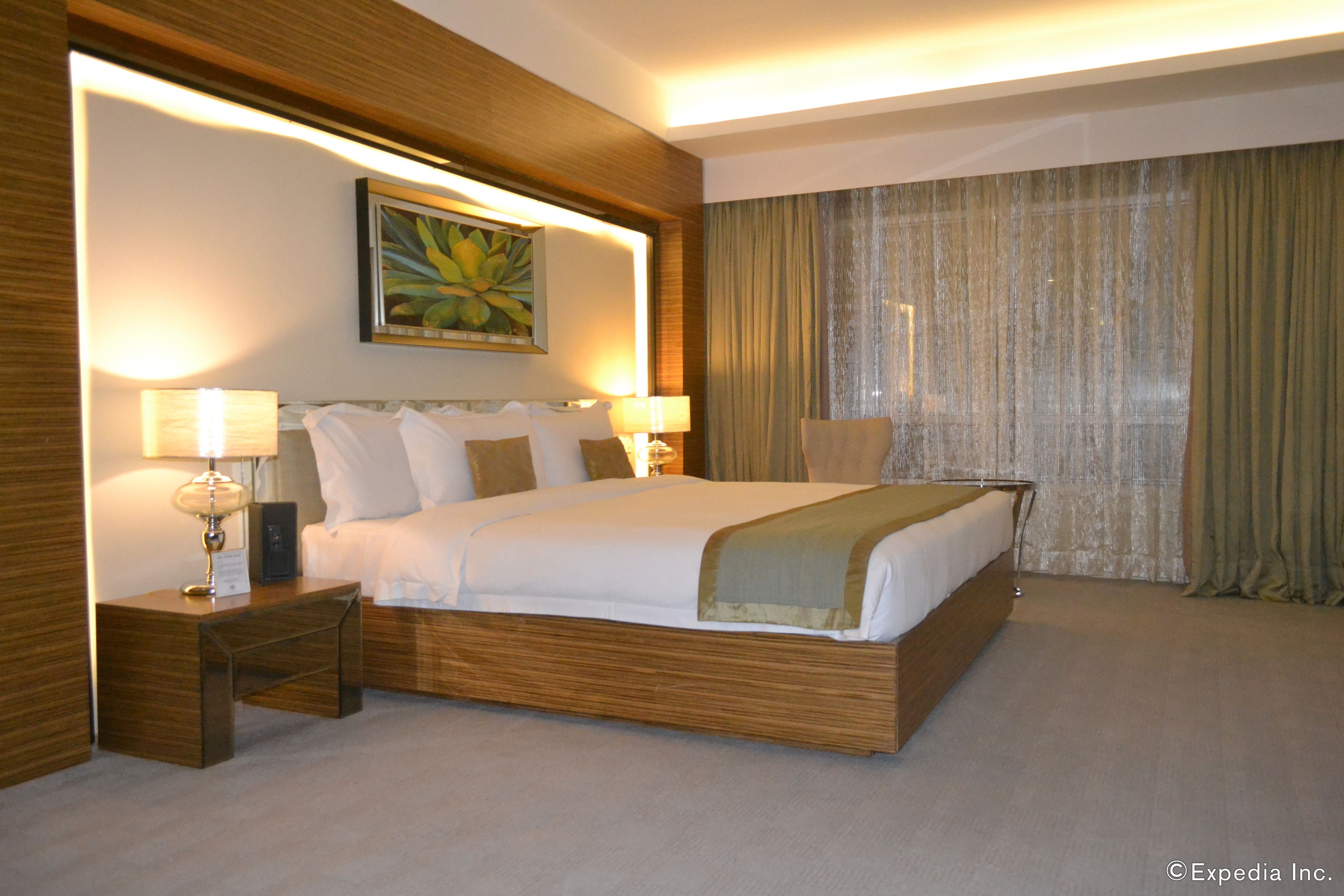 A hotel room with a large bed, two bedside tables with lamps, a painting above the bed, and a view of the outside through curtains.