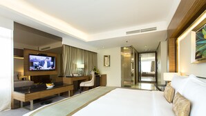 Una habitación de hotel moderna con una cama grande, un escritorio con televisor de pantalla plana, una zona de estar y un baño visible a través de una puerta abierta.