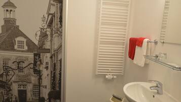 Un baño con lavamanos blanco, un espejo, un perchero para toallas y una ventana con contraventanas blancas.