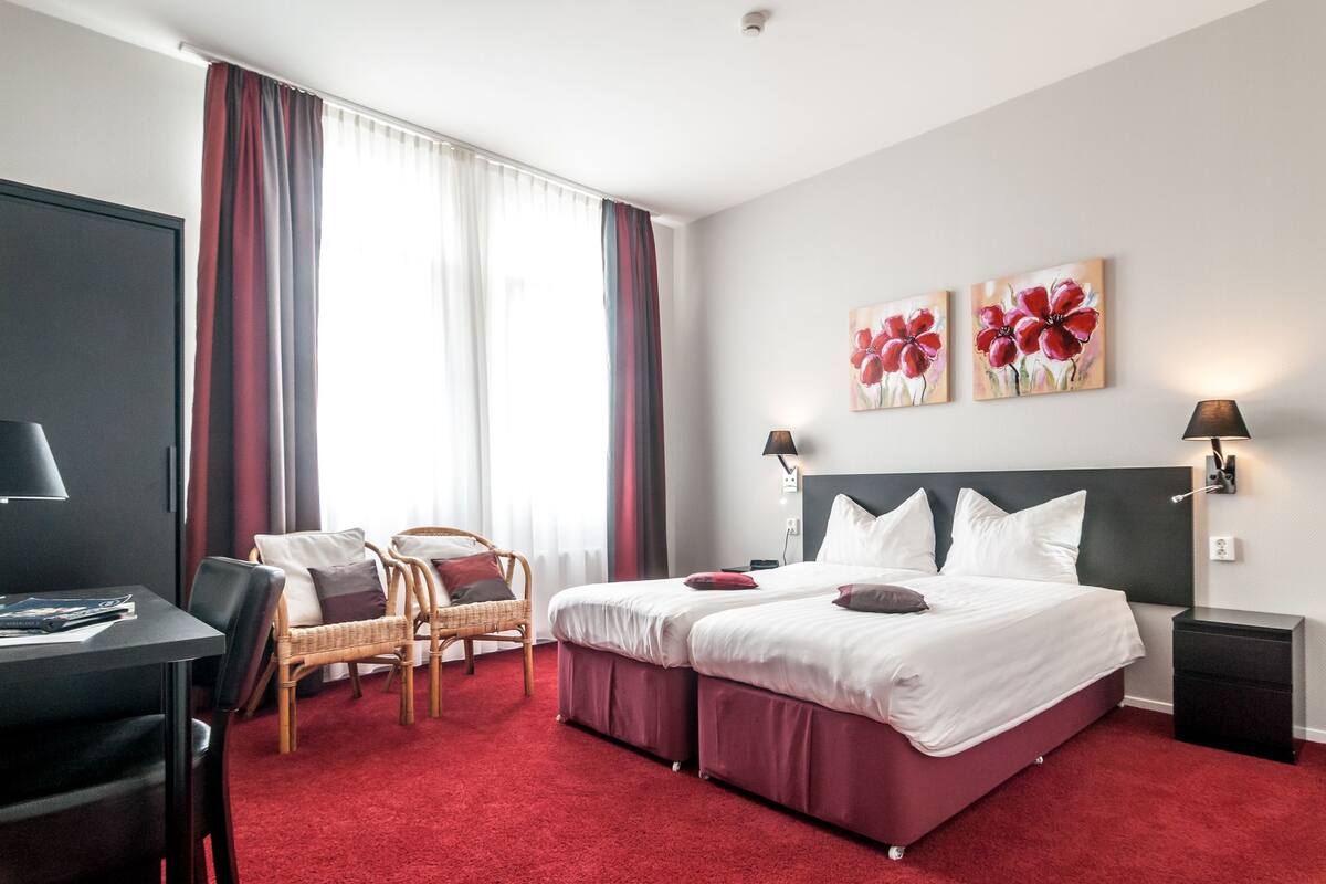 Une chambre d’hôtel avec un lit, deux tables de chevet, un bureau, une chaise et une moquette rouge.