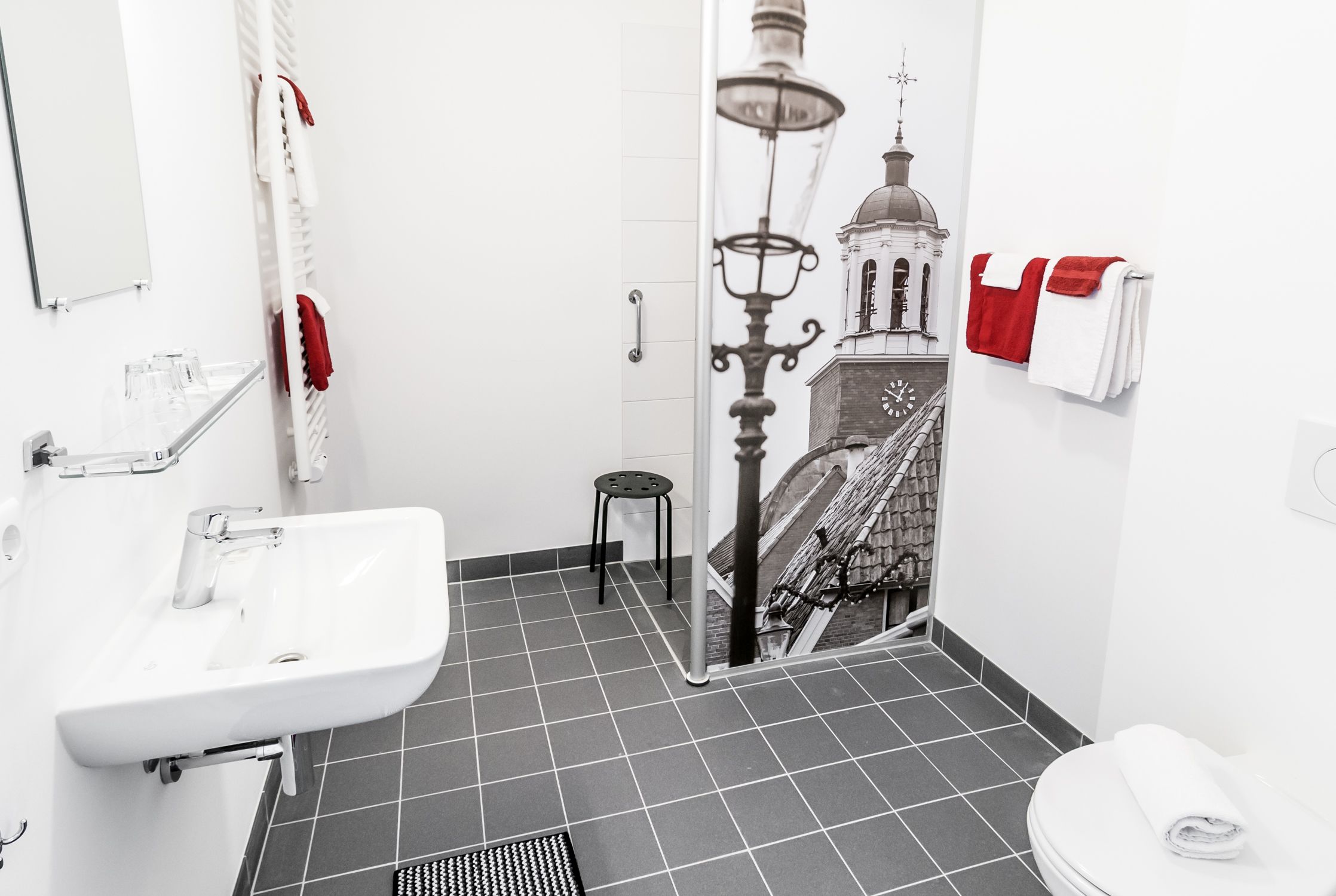 A bathroom with a white sink, a mirror, a towel rack with red towels, and a black stool.