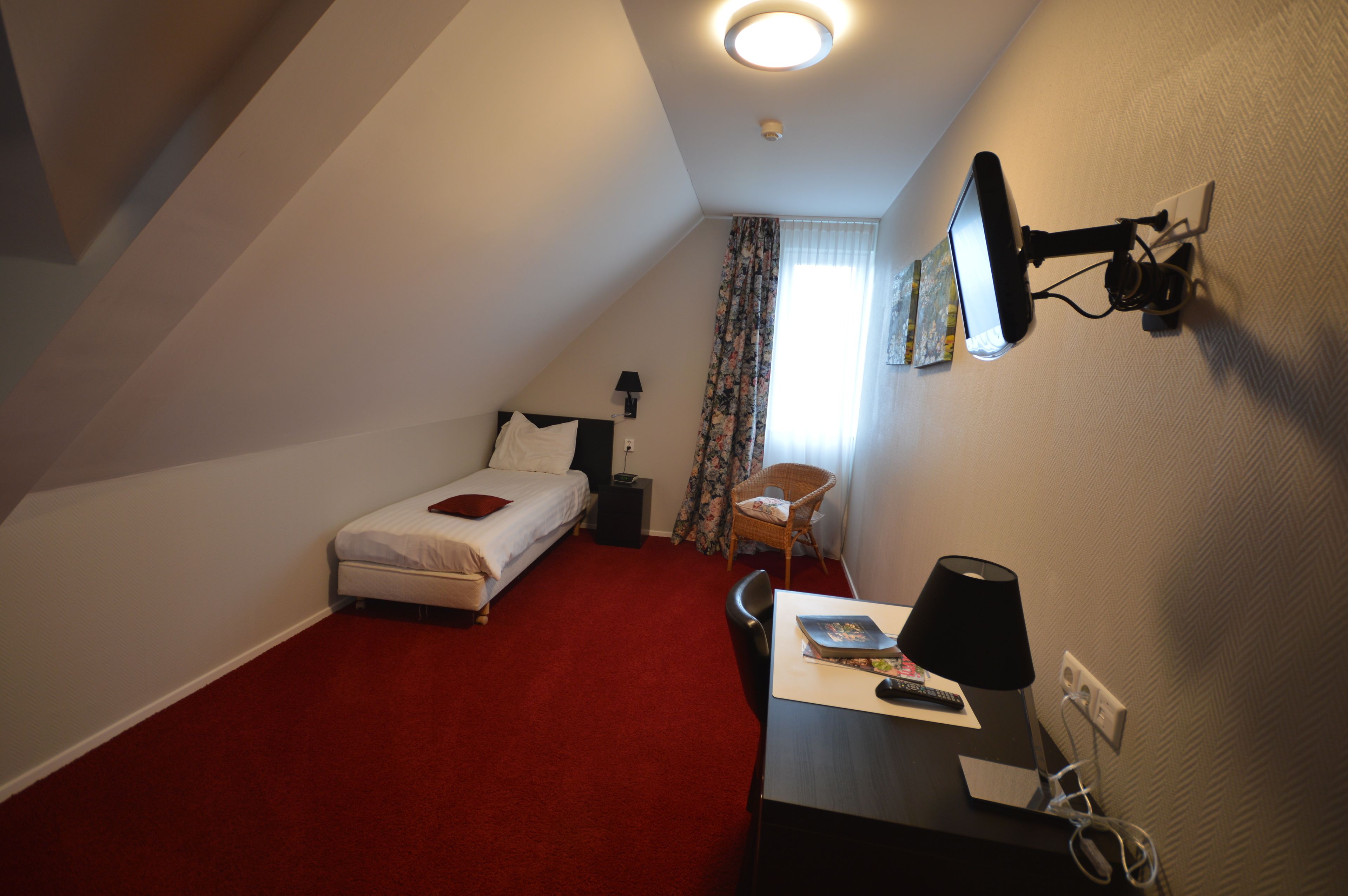 A small, triangular room with a bed, a desk, and a chair. There is a window with curtains, a lamp, and a wall-mounted TV.