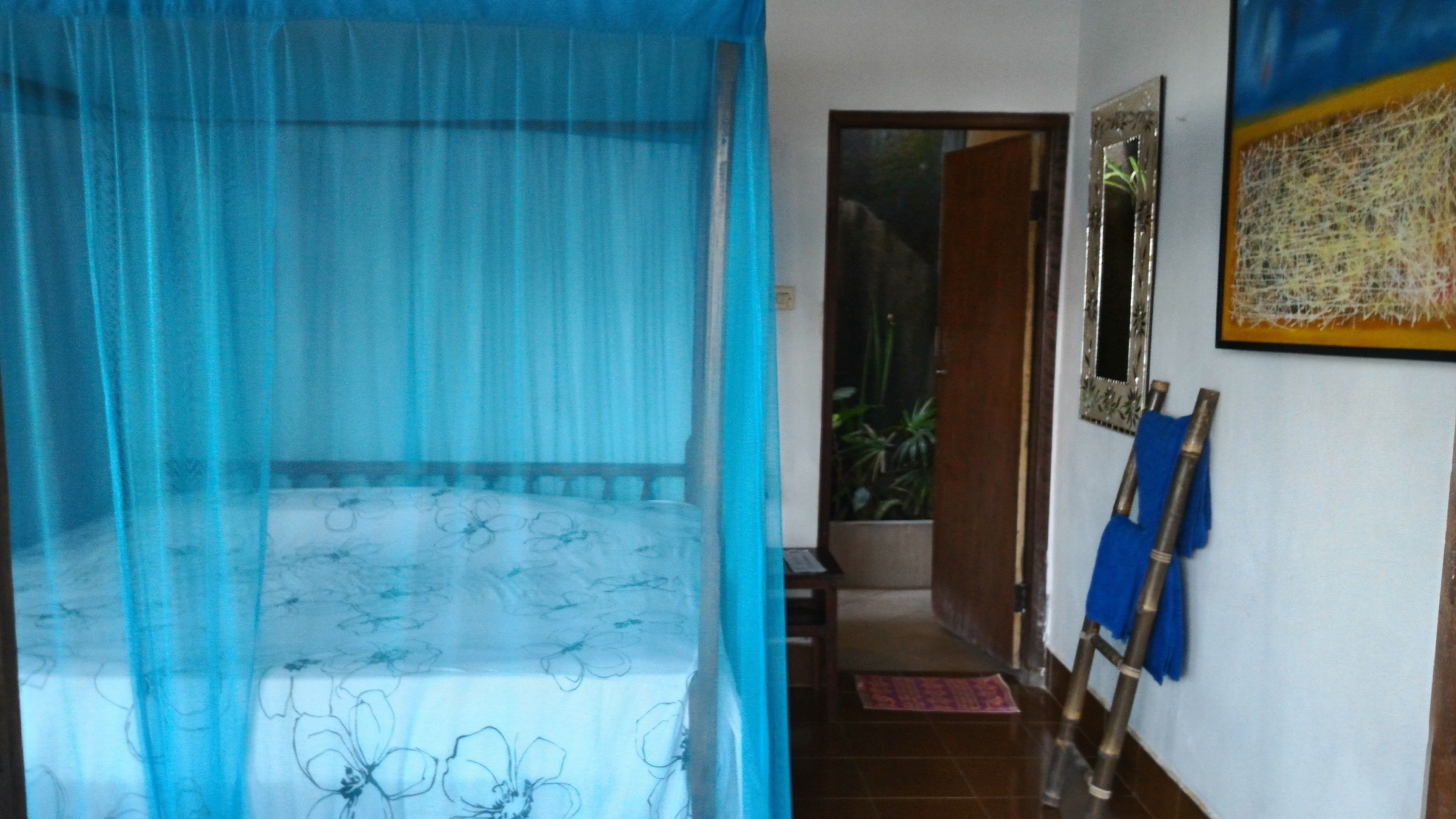 Una habitación con una cama cubierta por una cortina azul translúcida, una puerta de madera que da a otra habitación, y un espejo fijado a la pared.
