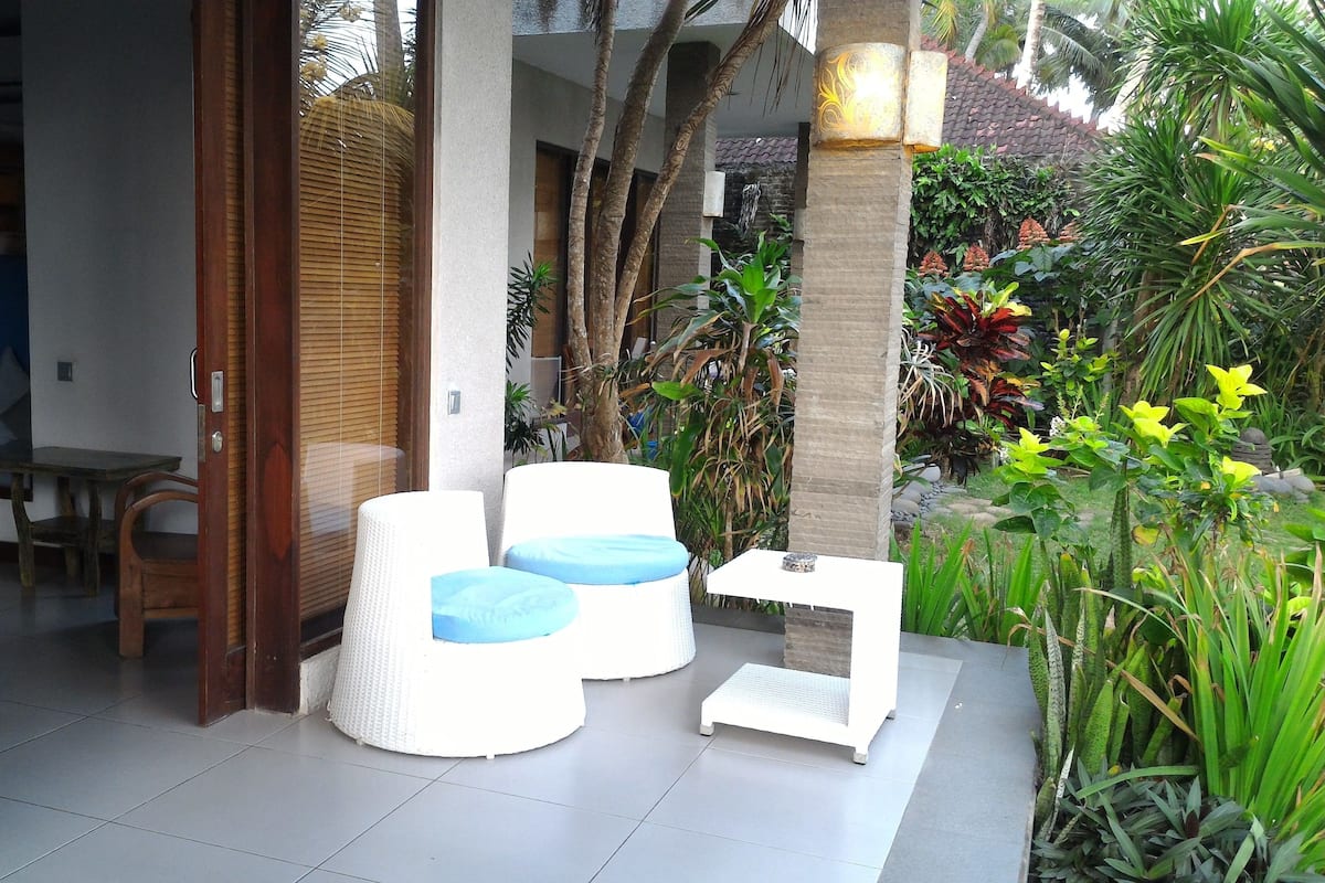 A modern outdoor seating area with two white chairs and a small table, surrounded by lush tropical plants and a stone wall.