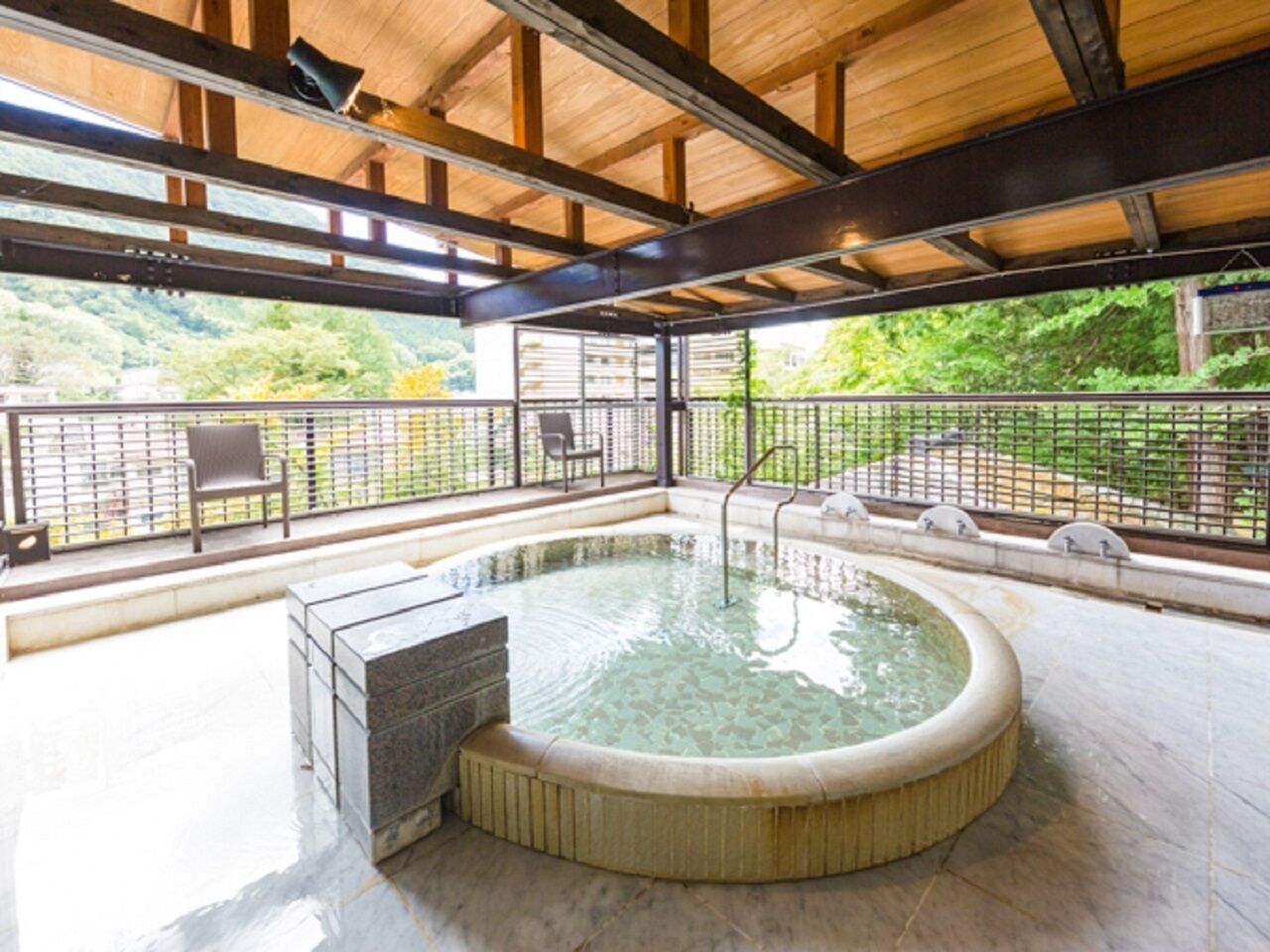 A hot tub with a wooden deck and metal railing, surrounded by a glass enclosure with a view of greenery.