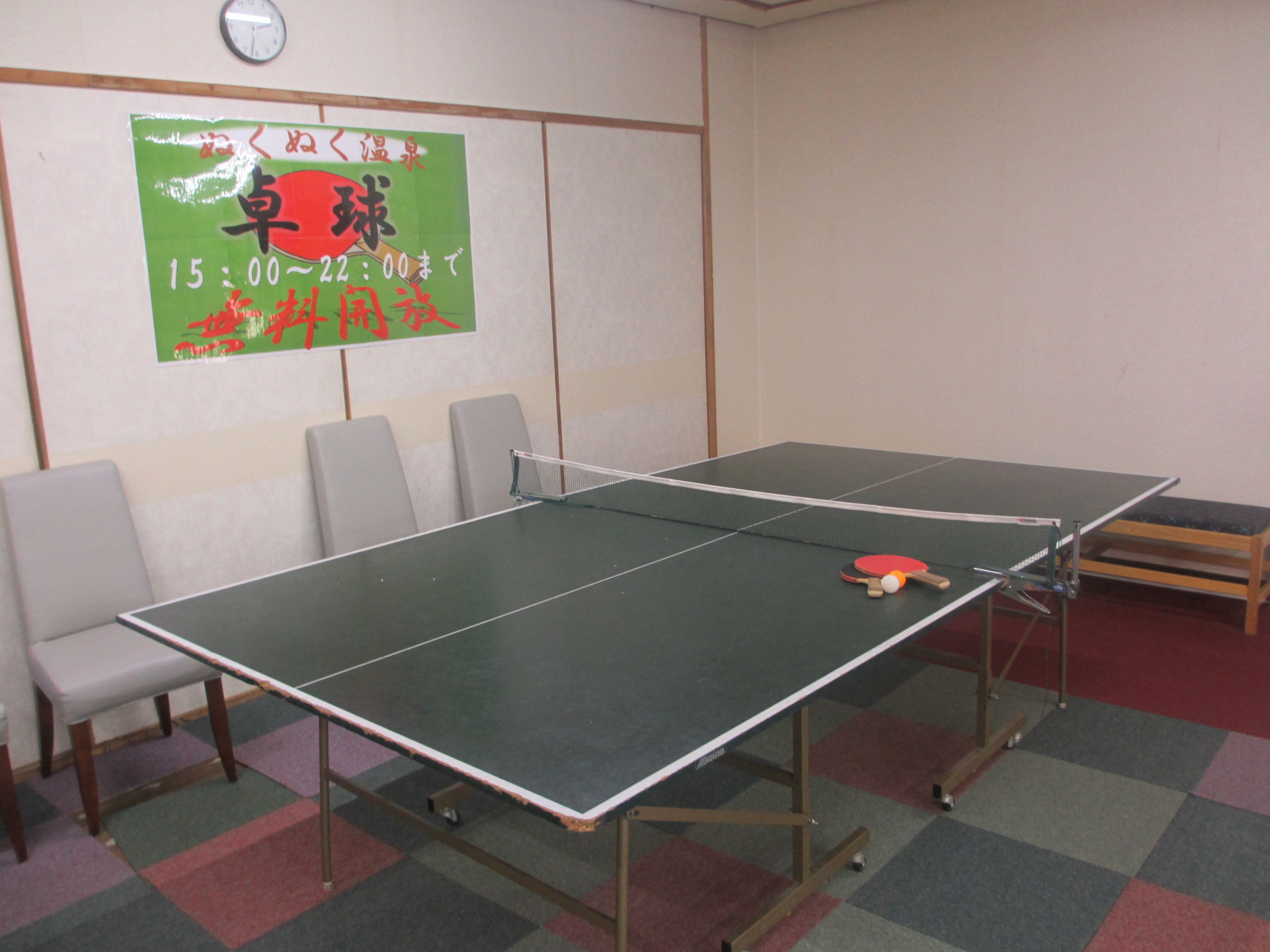 A ping pong table with chairs around it, a clock on the wall, and a poster with Japanese text.