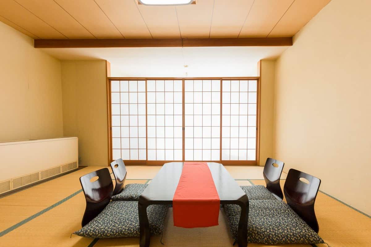 A traditional Japanese room with tatami flooring, a low table with a red runner, and four chairs with cut-out backs.