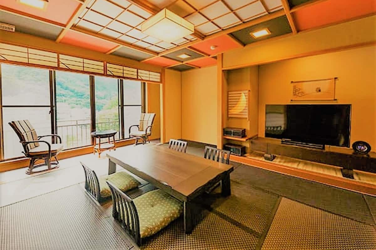A spacious room with a large dining table, chairs, a television, and a view of the outdoors through large windows.