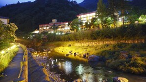 A riverside pathway with a wooden fence, a river with rocks, and buildings with illuminated windows in the background. - LiVEMAX RESORT KAWAJI (Nikko)