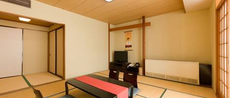 A traditional Japanese room with tatami flooring, a low table with a red runner, a television, and a futon bed.