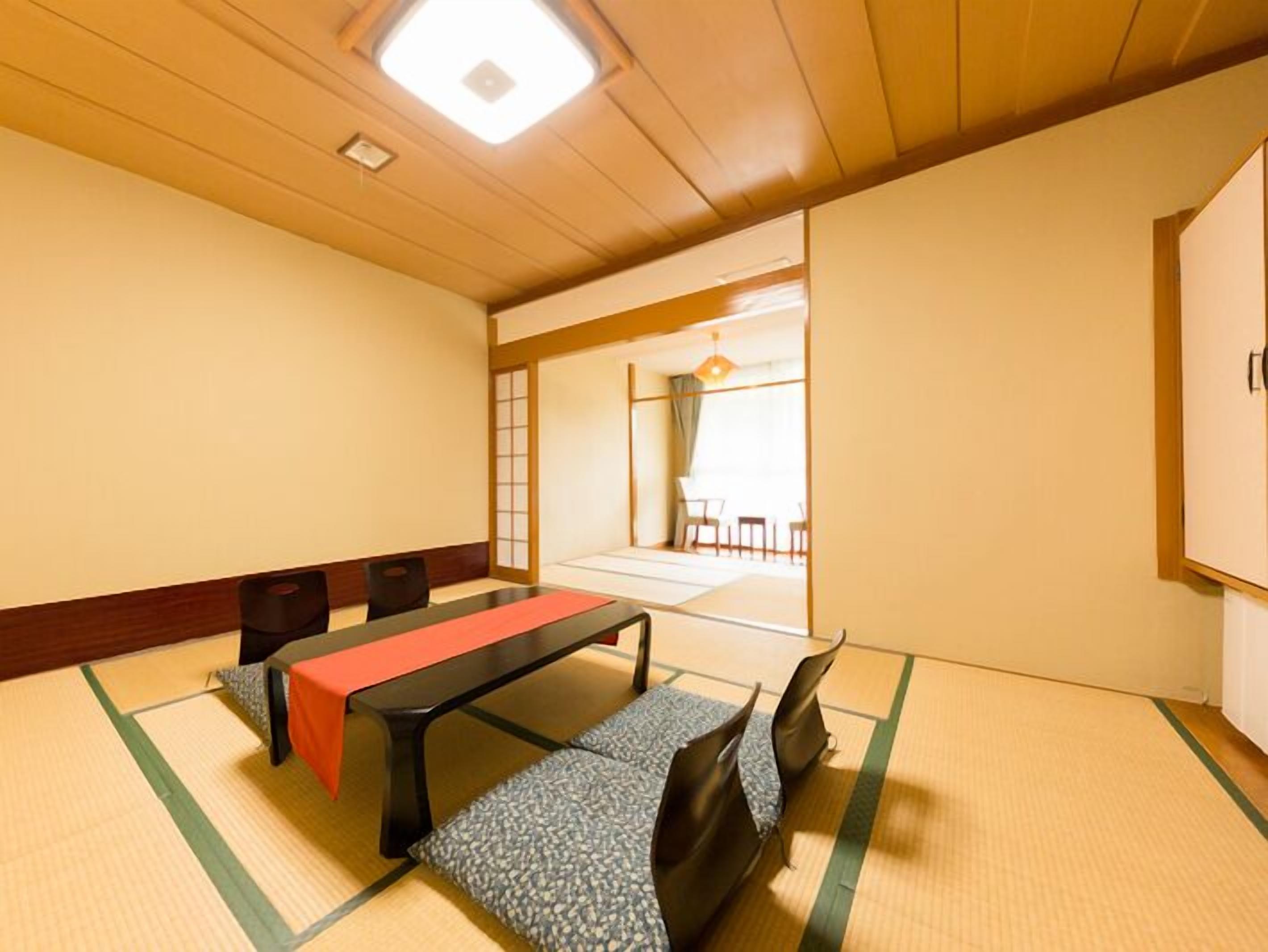 Ein Raum mit Tatami-Matten, einem niedrigen Tisch, Stühlen und einer verschiebbaren Tür zu einem anderen Raum.
