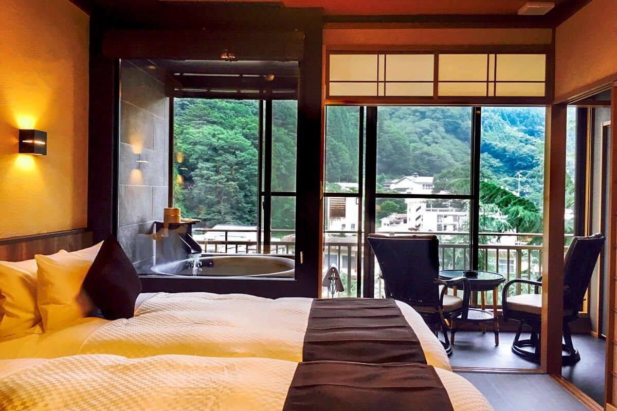A hotel room with a large bed, a view of the mountains, a balcony with chairs, and a bathtub.