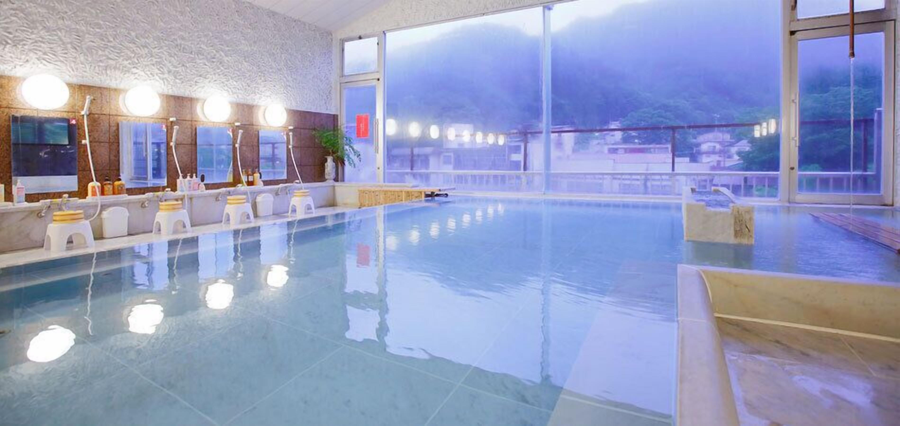 Una gran piscina interior con suelo de baldosas, una zona de estar con taburetes blancos y un amplio ventanal que ofrece vistas al exterior.