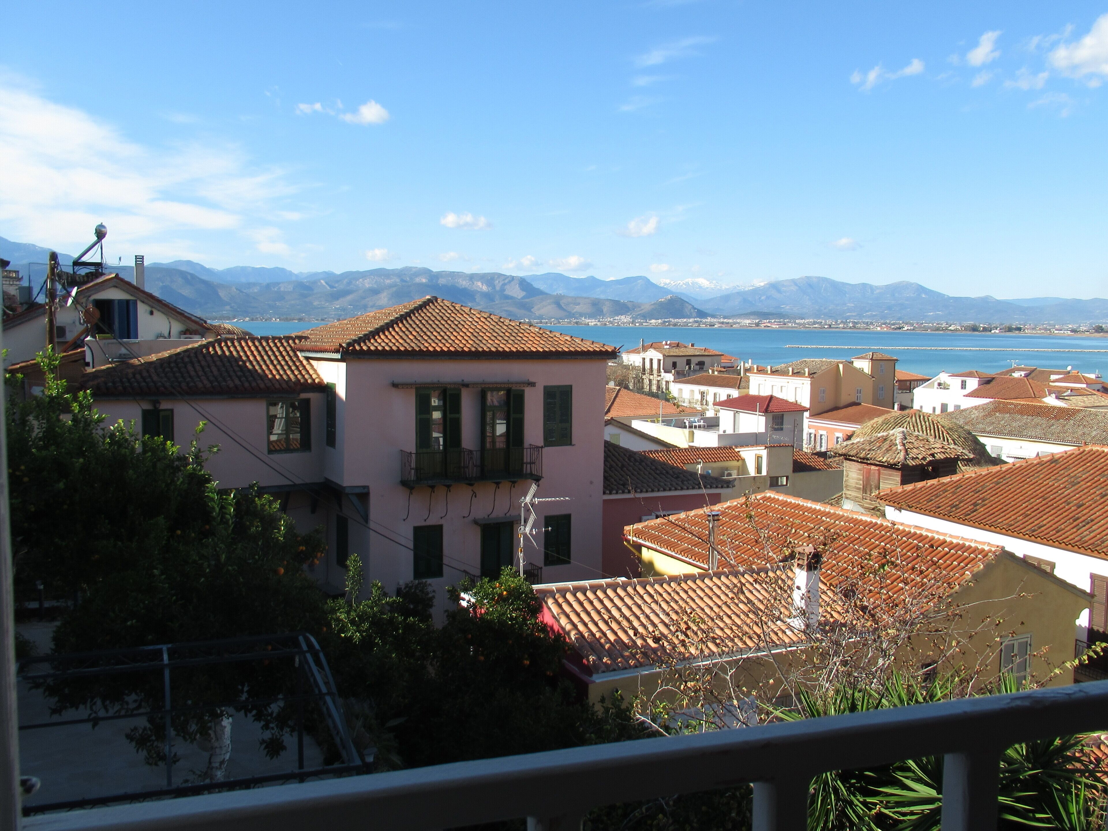 A coastal town with terracotta-roofed buildings, a clear blue sky, and a mountainous backdrop.