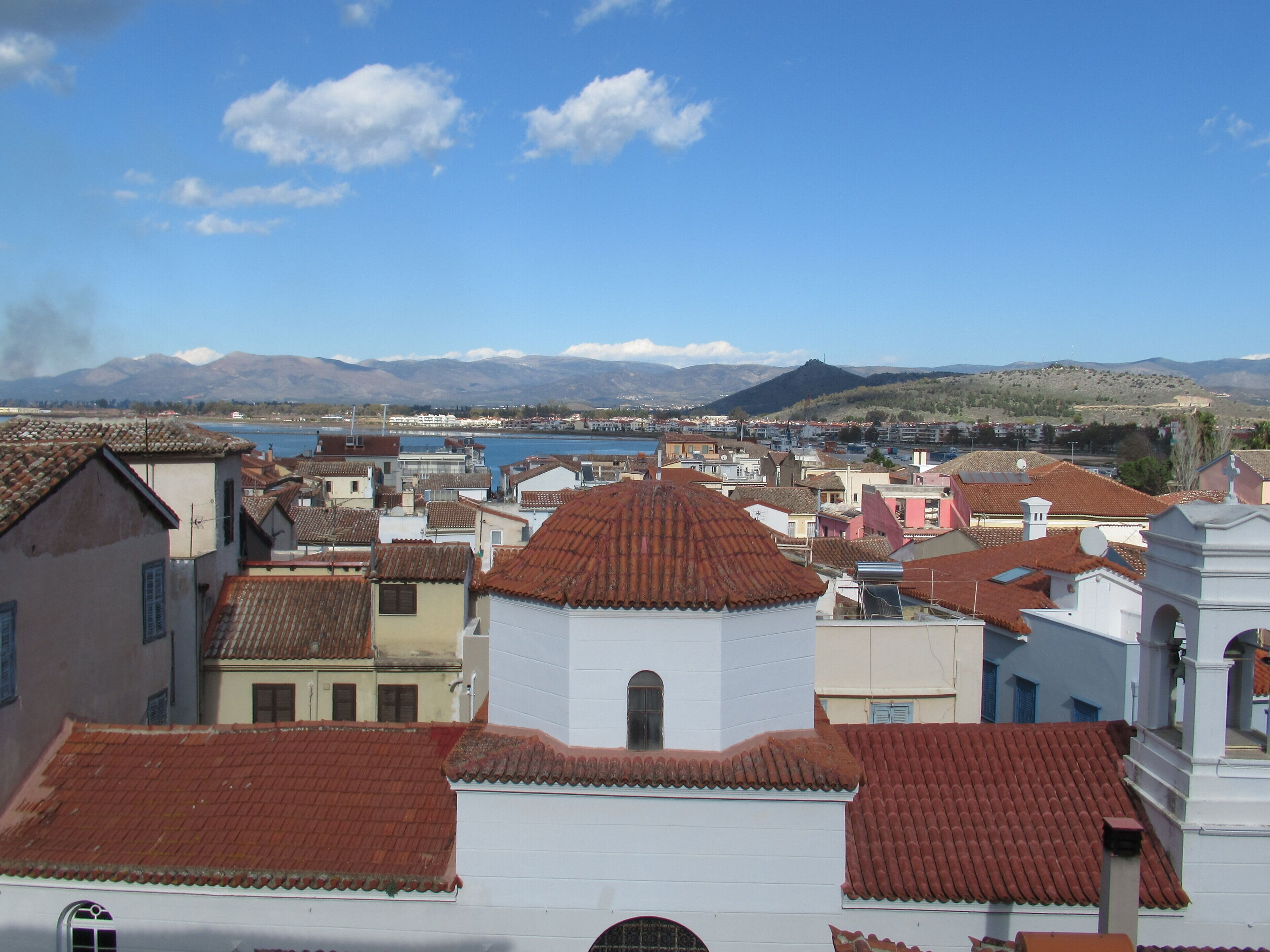 Un pueblo costero con edificios de tejados de terracota, una iglesia con campanario y una cadena montañosa al fondo.
