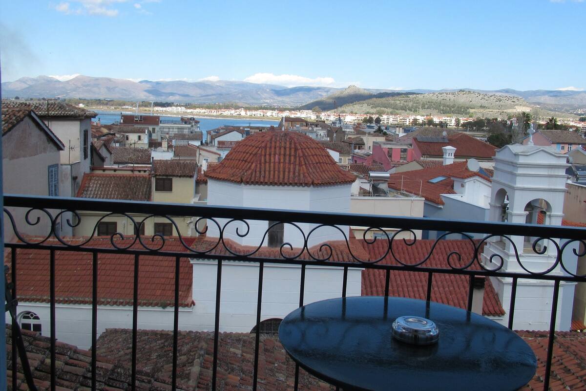 A balcony with a view of a town, including buildings with red roofs and a body of water in the distance.