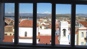 A view through a window with a metal frame, featuring a town with red-tiled roofs, white buildings, and mountains in the background. - Byron Hotel (Nafplio)