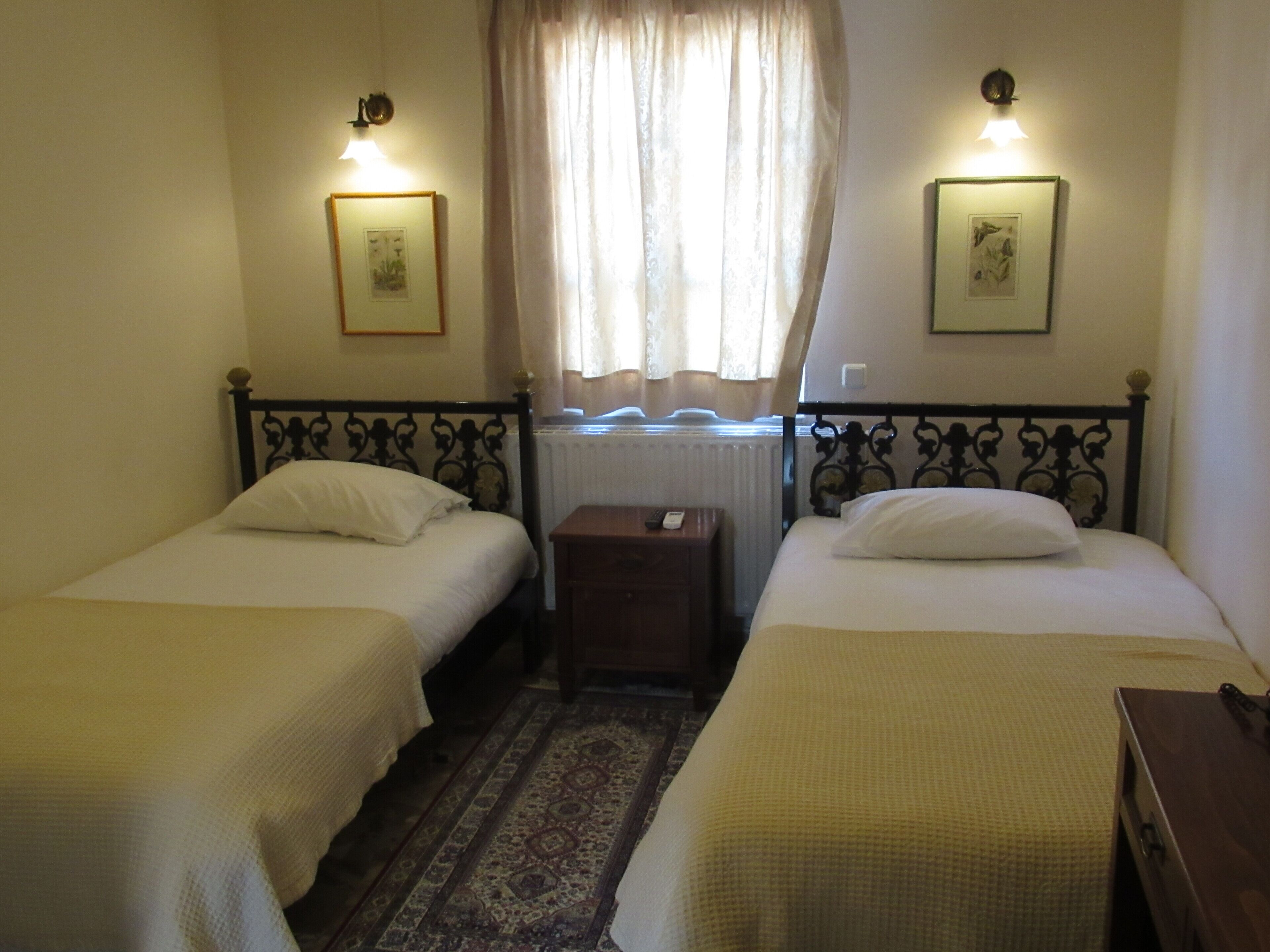Quarto com duas camas de solteiro, pequena mesa de cabeceira, luminárias fixadas na parede e uma janela com cortinas.