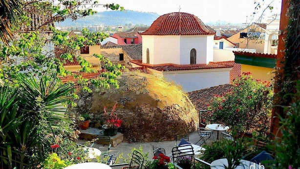 A courtyard with outdoor seating, a large stone structure, and buildings in the background. - Byron Hotel (Nafplio)