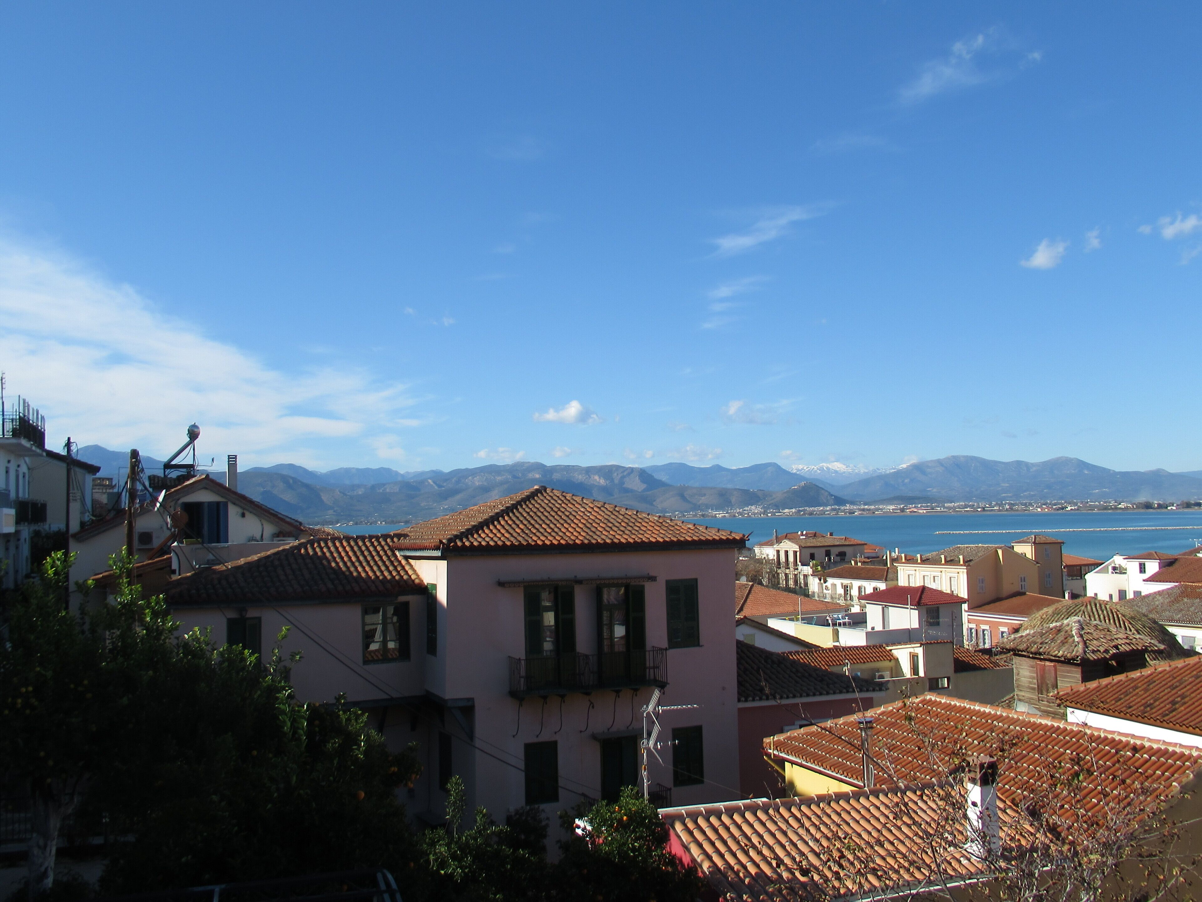 A coastal town with terracotta-tiled buildings, a clear blue sky, and a mountainous backdrop.