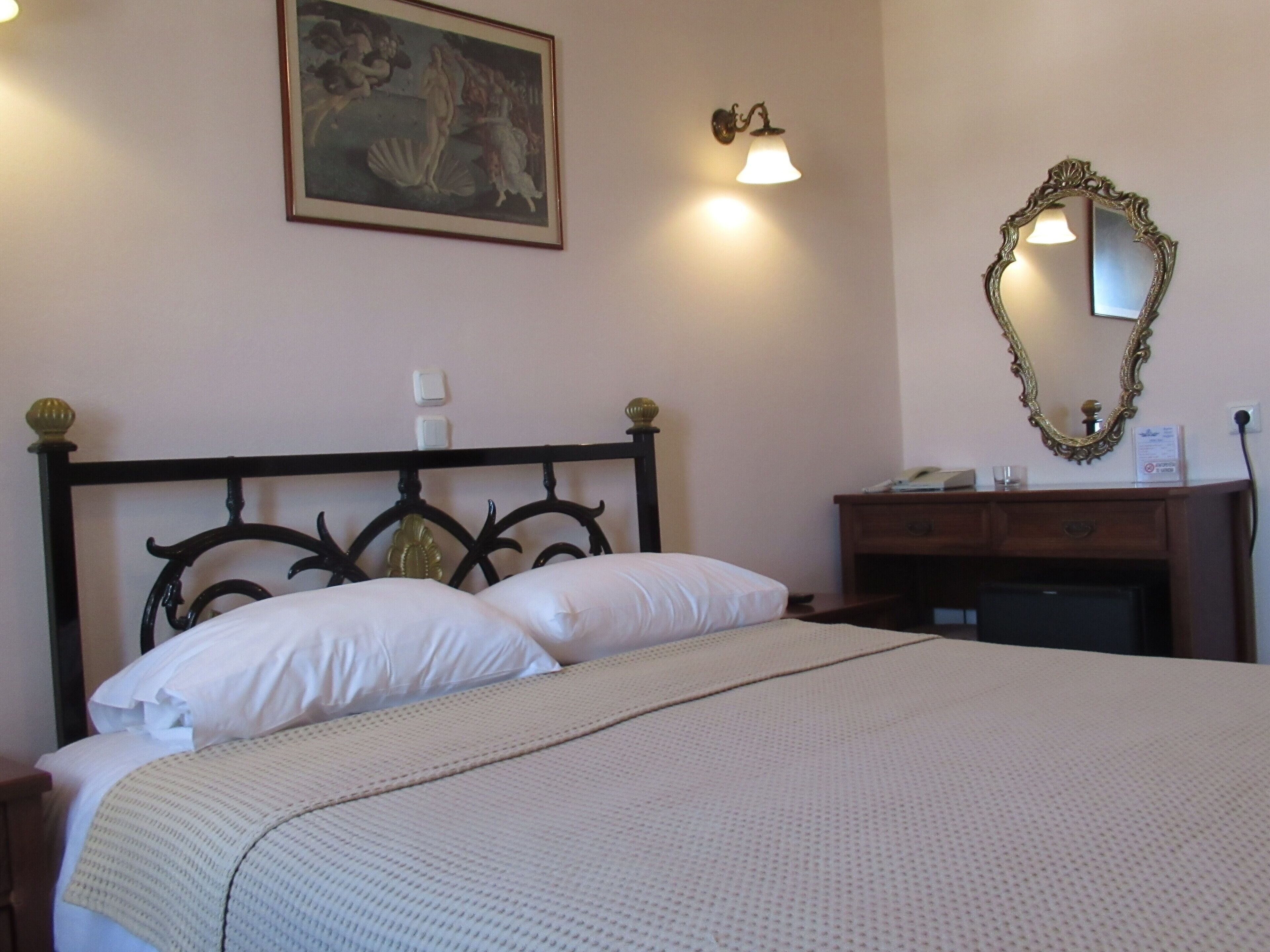 Un lit bien fait, avec du linge de lit blanc, une tête de lit en bois, une table de chevet avec une lampe, une commode et un miroir fixé au mur.