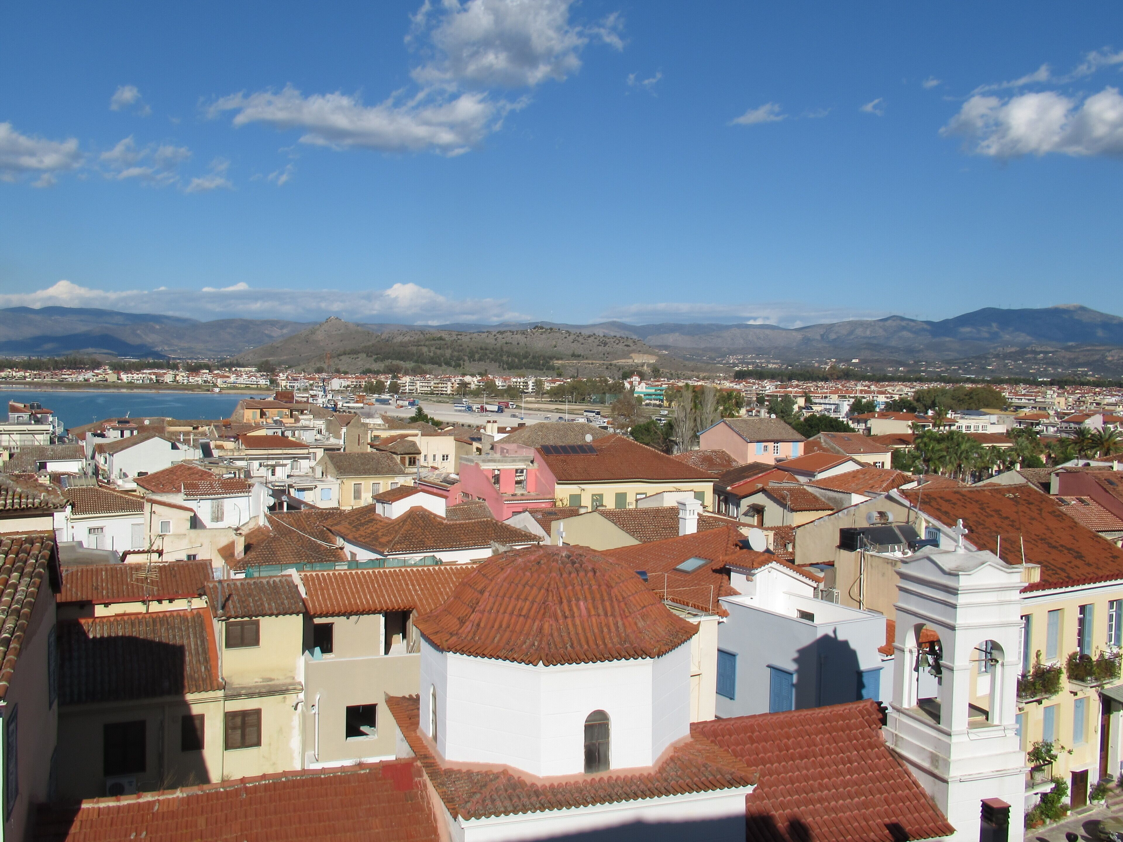 A coastal town with terracotta-roofed buildings, a prominent white church, and a view of the sea and mountains in the distance.