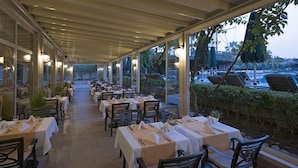 Un comedor interior con mesas dispuestas para huéspedes, amplias ventanas con vistas a una zona de piscina y un paseo cubierto con sillas.