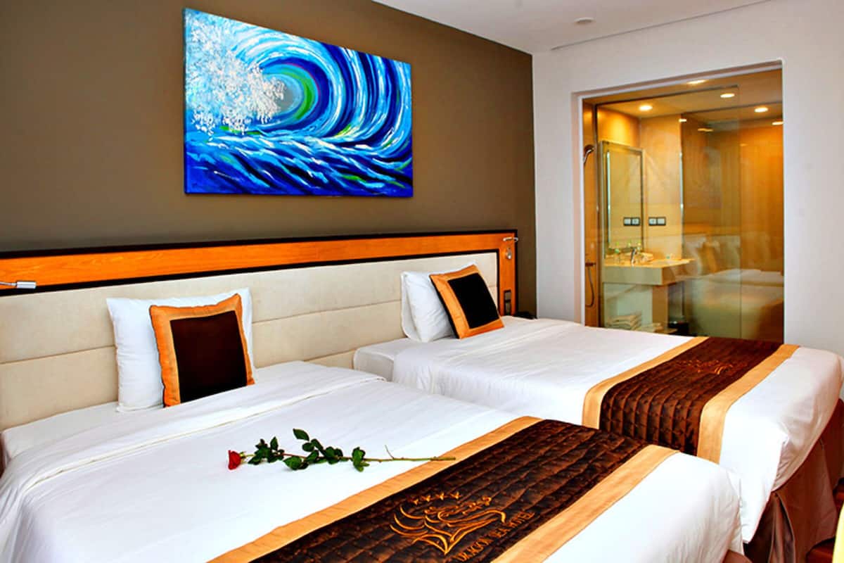 A hotel room with two beds, a large painting, and a bathroom visible through a glass door.