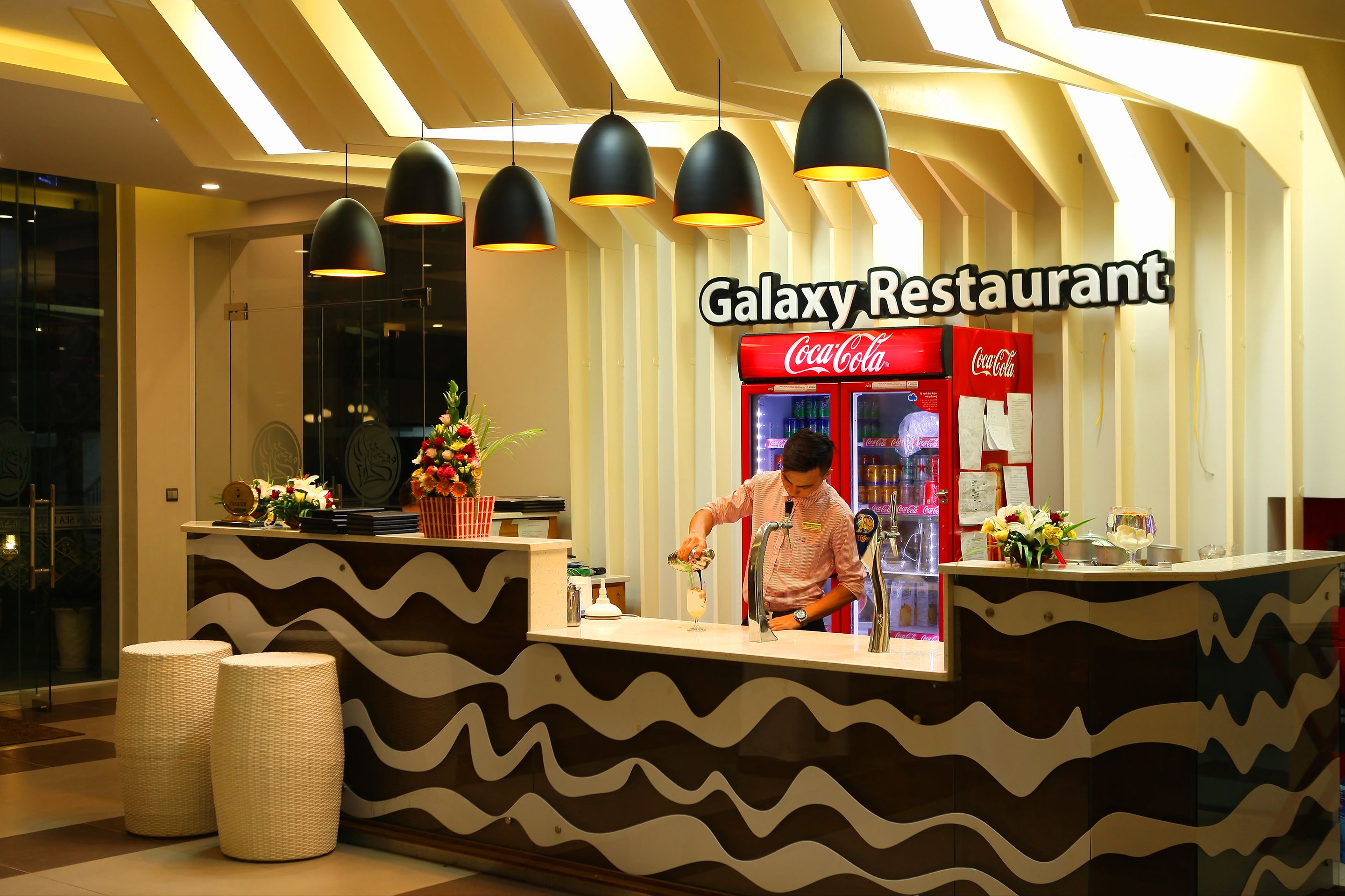 Un restaurant futuriste avec un réfrigérateur Coca-Cola, un comptoir au design ondulé et une personne en train de préparer une boisson.