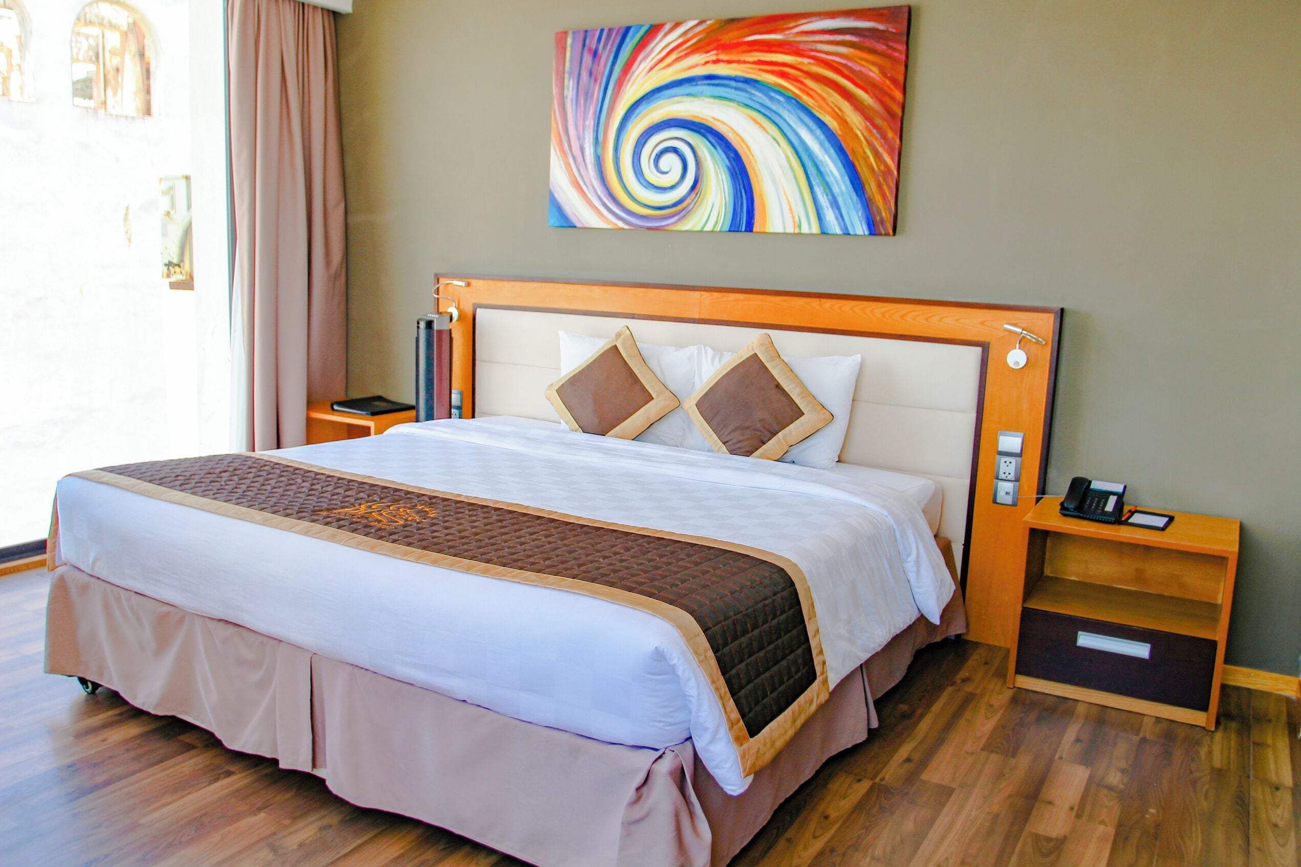 A hotel room with a bed, a nightstand, a phone, a wall art piece, and a window with curtains.