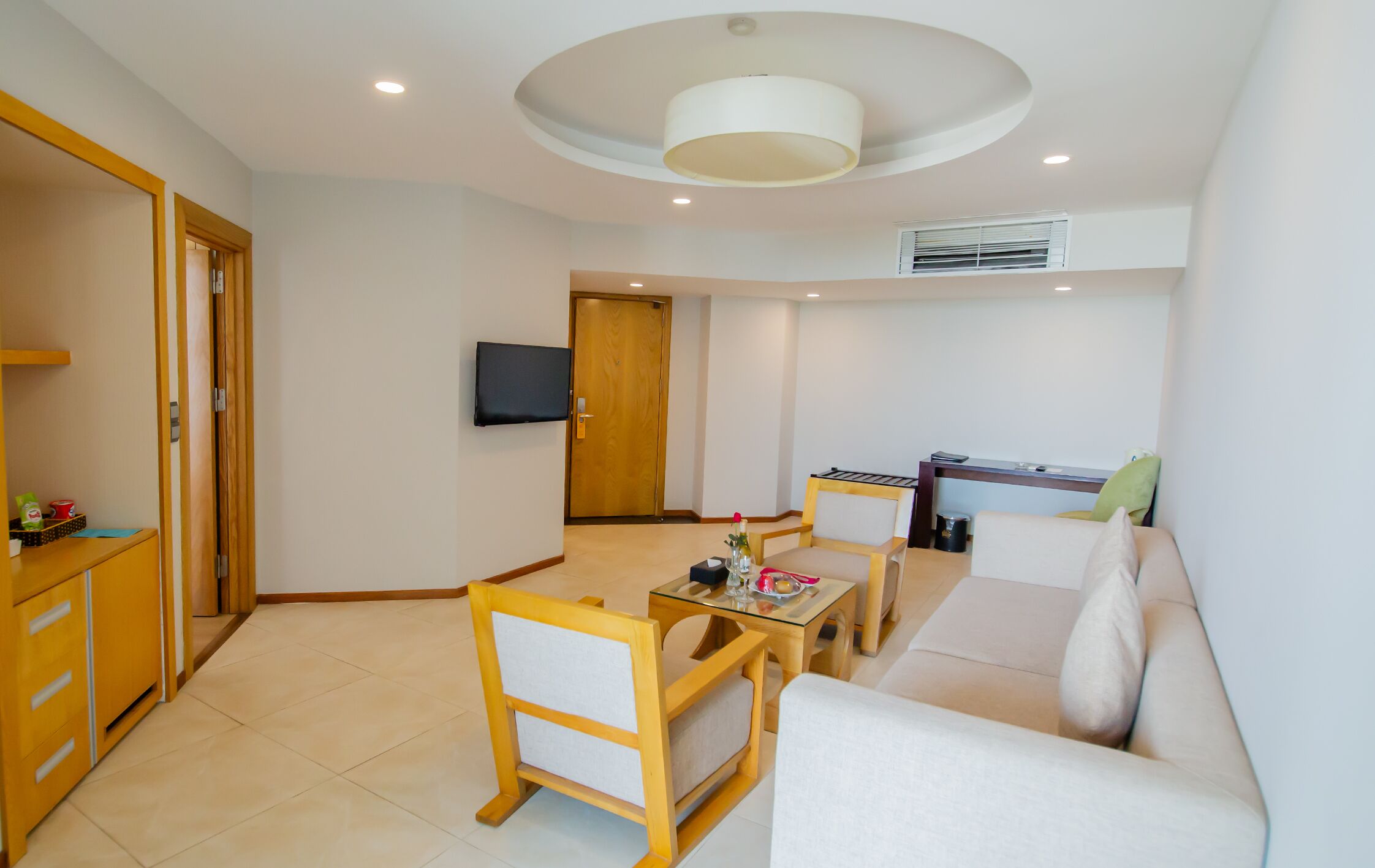 A modern living room with a sofa, a dining table with chairs, a TV mounted on the wall, and a kitchen area with a stove.