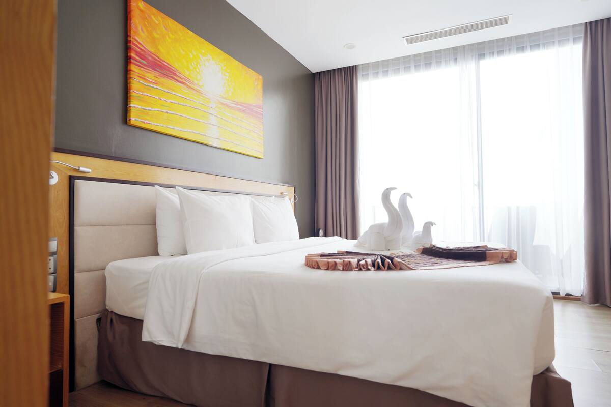 A hotel room with a bed, a painting, a large window, and a view of the sky.