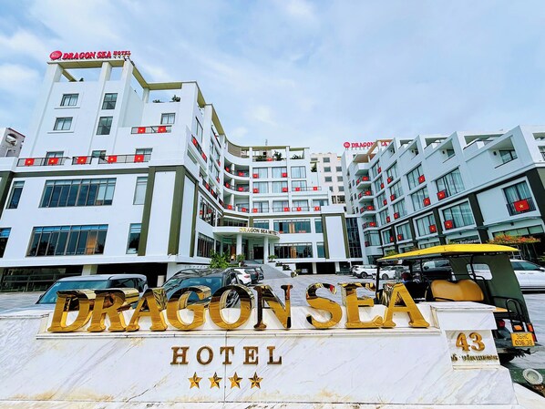 A multi-story hotel with a prominent 'Dragon Sea Hotel' sign, multiple balconies with red flags, and a yellow vehicle in front. - Dragon Sea Sam Son Hotel (Sam Son)