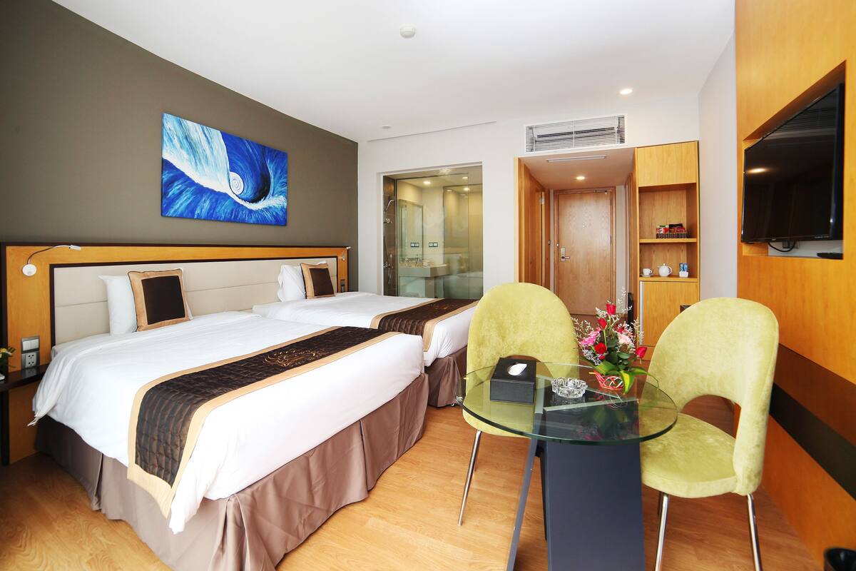 A hotel room with two beds, a glass table with chairs, a painting on the wall, and a TV mounted on the wall.