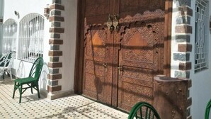 A large wooden door with intricate carvings, a white wall with a window, and a tiled patio with chairs. - Malabata Guest House (Tangier)