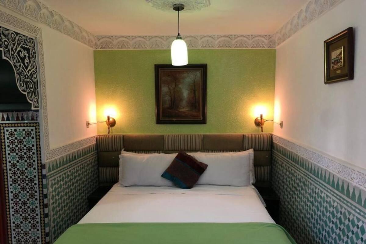 Habitación de hotel con cama, paredes verdes, detalles de baldosas estampadas y dos lámparas de pared.