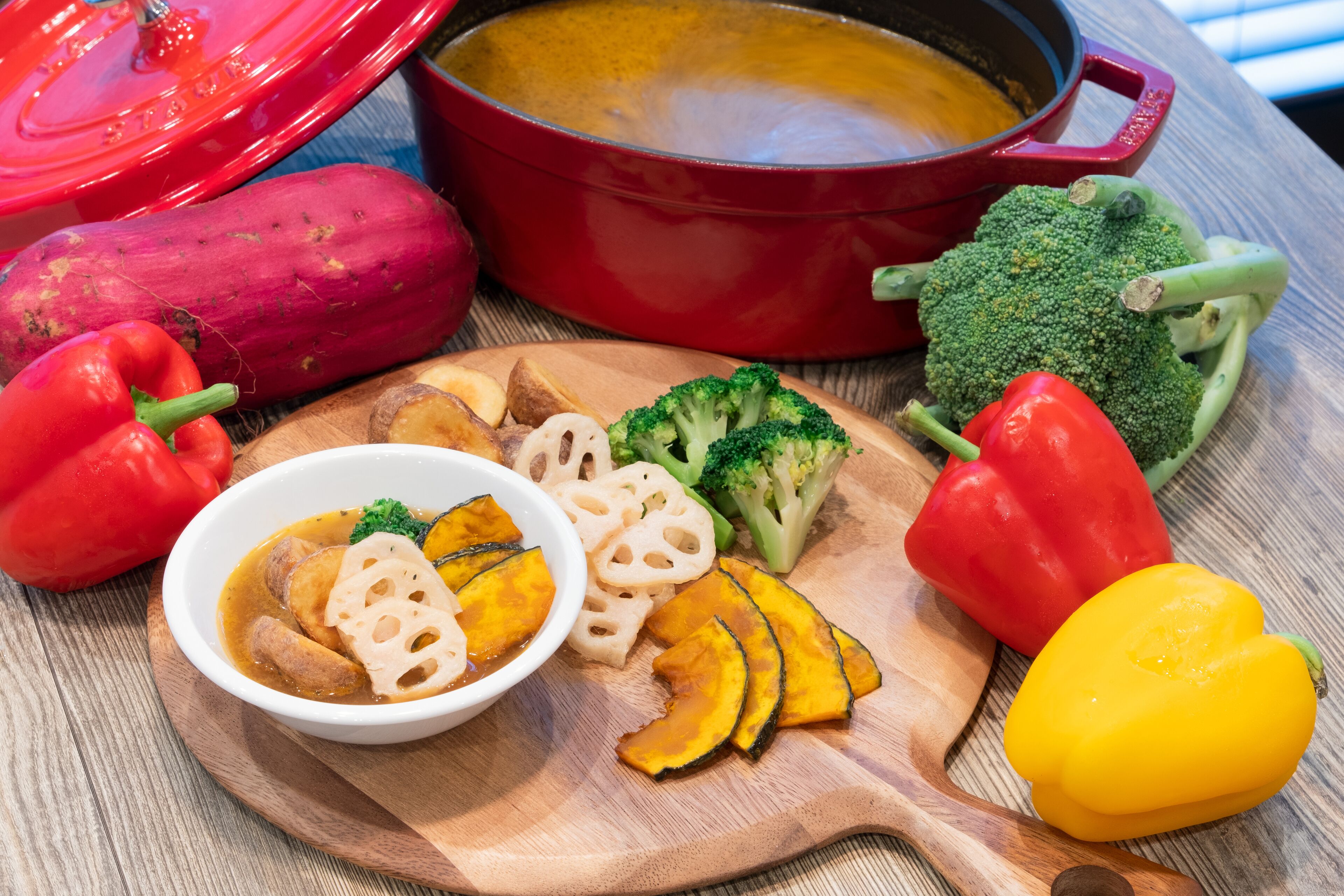 Taula de fusta amb un bol de sopa, verdures tallades i una olla vermella.