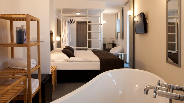 Una habitación de hotel moderna con una bañera grande, estantes de madera con toallas, una cama bien hecha y un televisor montado en la pared.