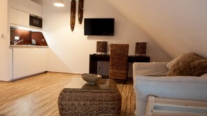 Una sala de estar moderna con un sofá, un televisor montado en la pared, una zona de cocina con armarios y un microondas, y piso de madera.