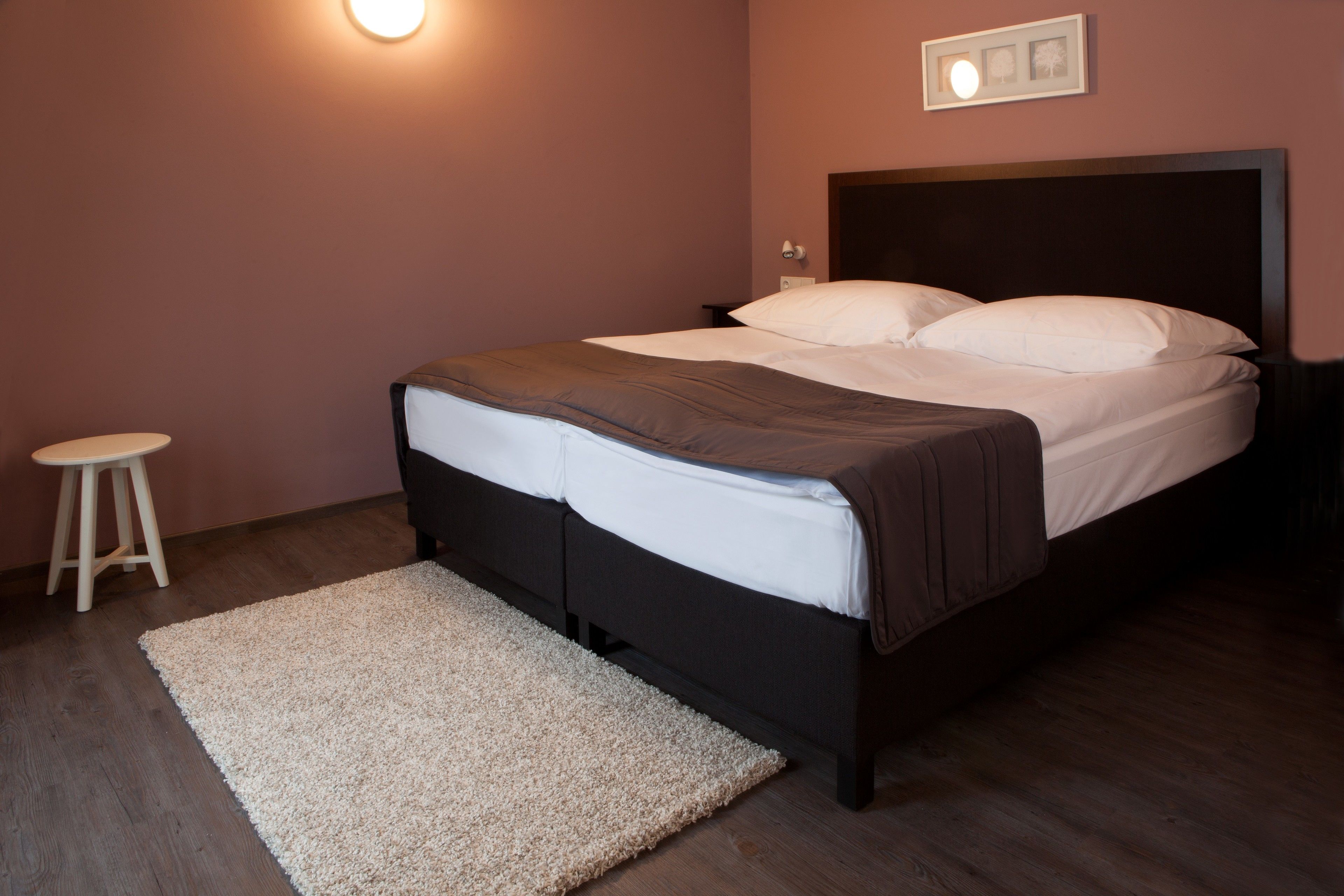 A neatly made bed with white and brown bedding, a small wooden stool, and a light-colored rug on a wooden floor.
