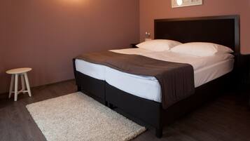 A neatly made bed with white and brown bedding, a small wooden stool, and a light-colored rug on a wooden floor.