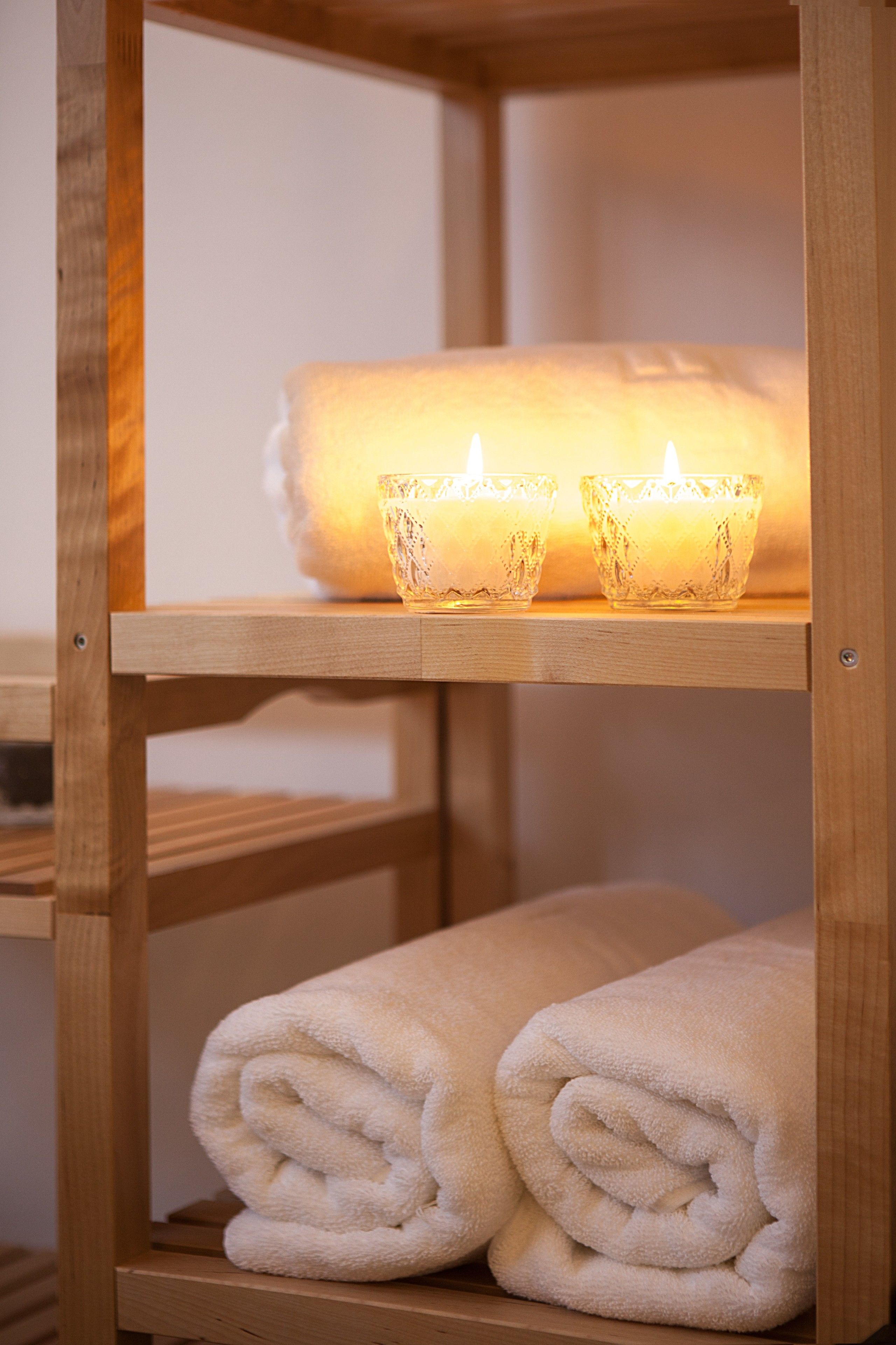 A wooden shelf with a rolled white towel and two lit candles in glass holders.