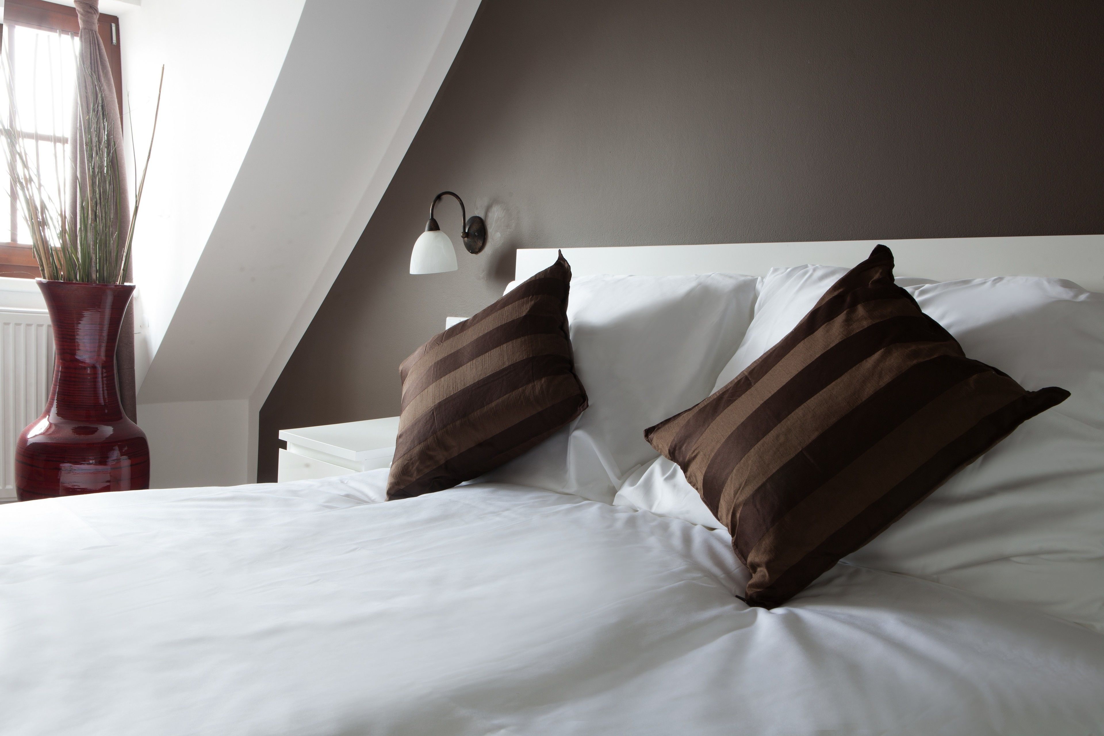 A neatly made bed with white linens and two striped pillows, a red vase with a plant, and a wall-mounted lamp.