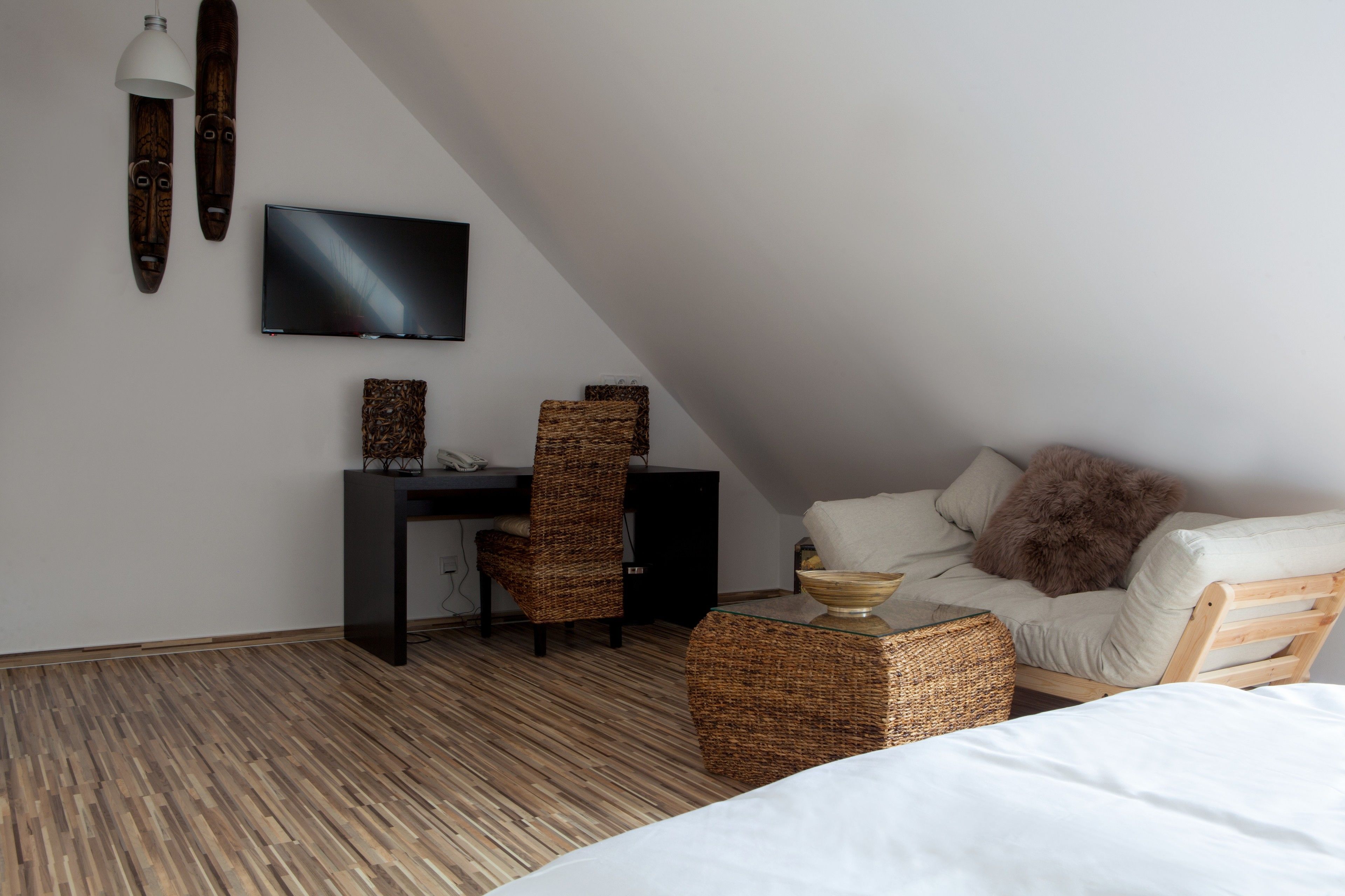 Ett modernt, väl upplyst rum med ett sluttande tak, en platt-TV som är monterad på väggen, ett skrivbord i trä med en stol, en bekväm soffa med en fluffig kudde och korgar av rotting på golvet.