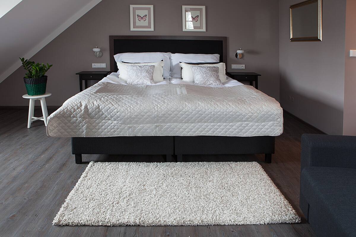 A neatly made bed with a quilted duvet, a white rug, and a small potted plant on a white stool.