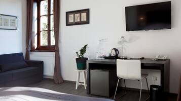 A hotel room with a bed, a desk with a computer, a TV, a small fridge, a chair, a potted plant, and a window with curtains.