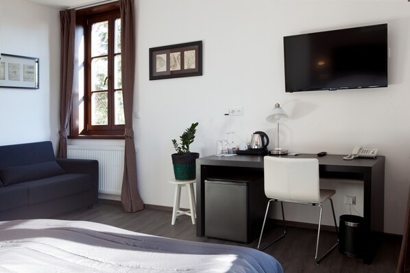Een hotelkamer met een bed, bureau met computer, televisie, kleine koelkast, stoel, potplant en raam met gordijnen.