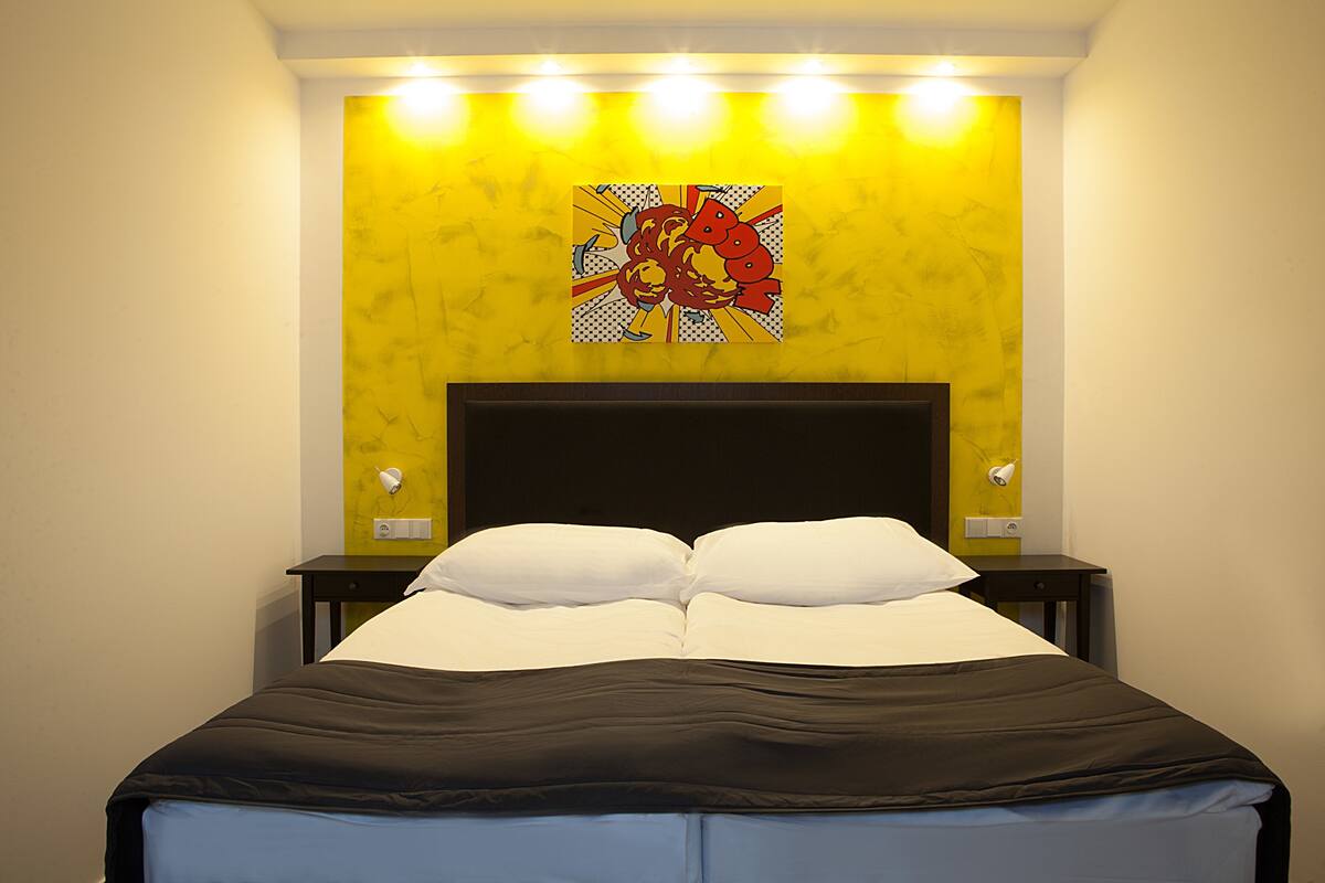 A bed with white and black bedding, a yellow accent wall, and a red and yellow artwork above it.