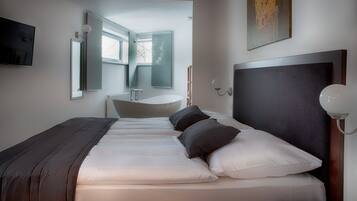 A modern hotel room with a large bed, a bedside table, a nightstand, a wall-mounted TV, and a window with an open door leading to a bathroom.